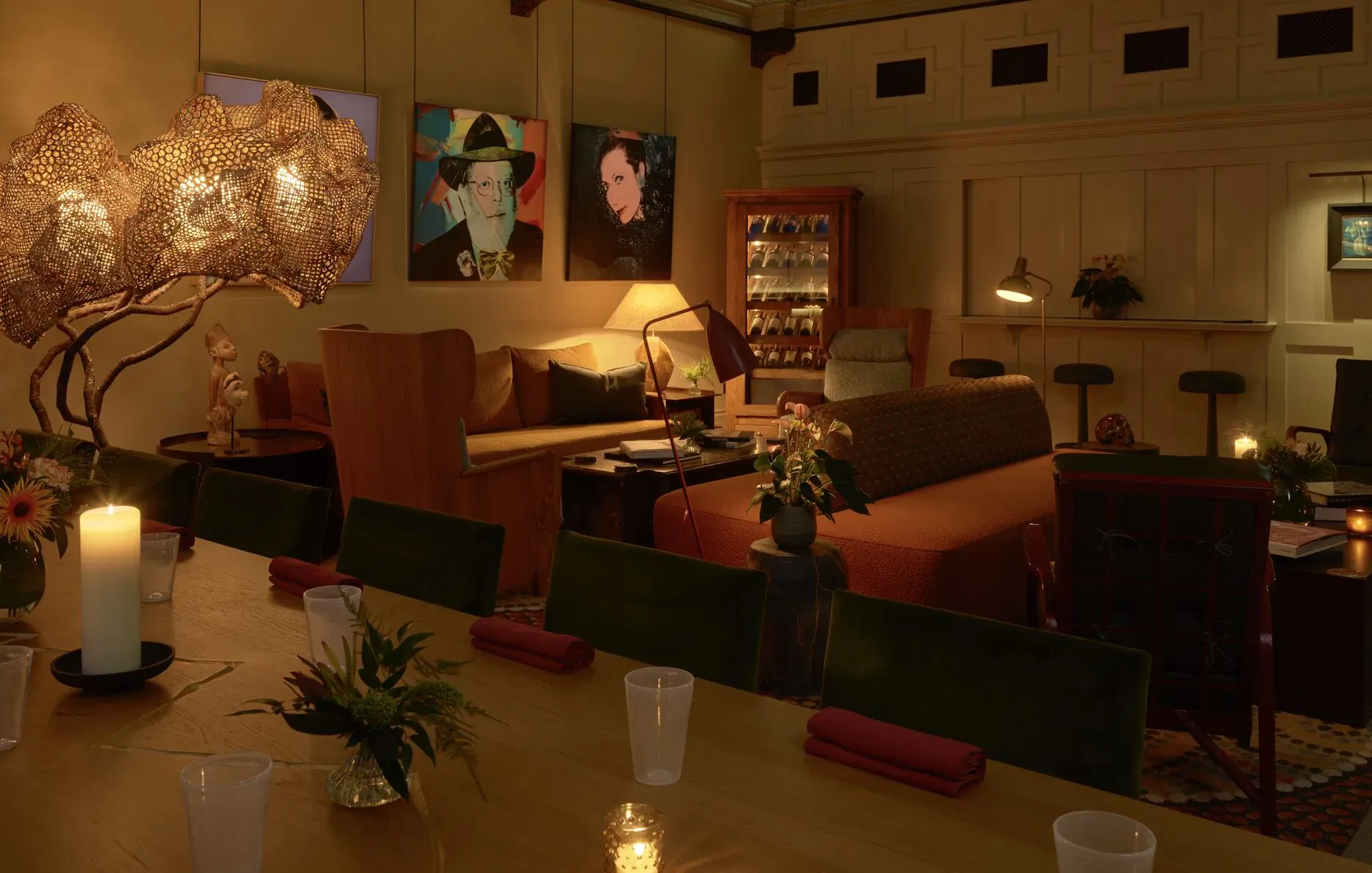 Cozy dimly lit dining room with candlelit wooden table, plush chairs, Warhol-style art, and vintage bar cabinet.
