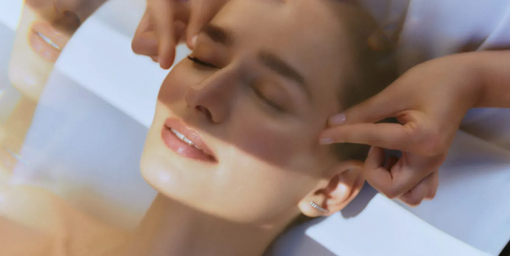 Close-up of woman receiving facial massage at spa, eyes closed, hands gently pressing temples on white surface