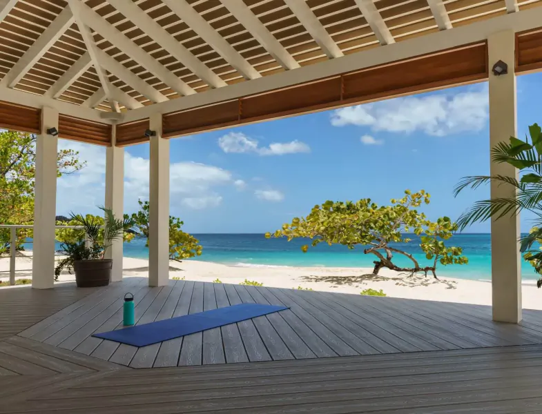 Open wooden yoga pavilion at Spice Island Beach Resort, blue yoga mat and water bottle on deck, turquoise sea and beach view.