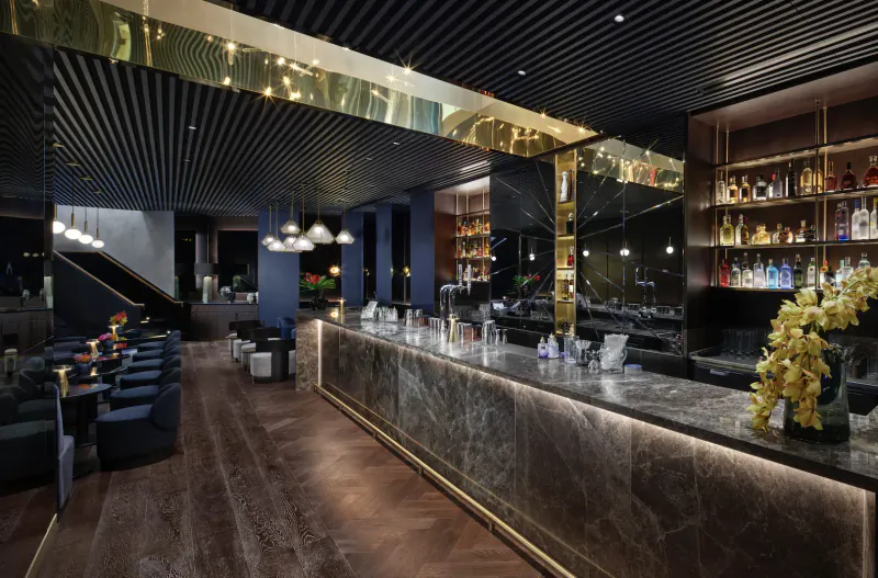 Luxurious modern hotel bar with marble counter, gold accents, wooden floors, blue seating, and yellow flowers.