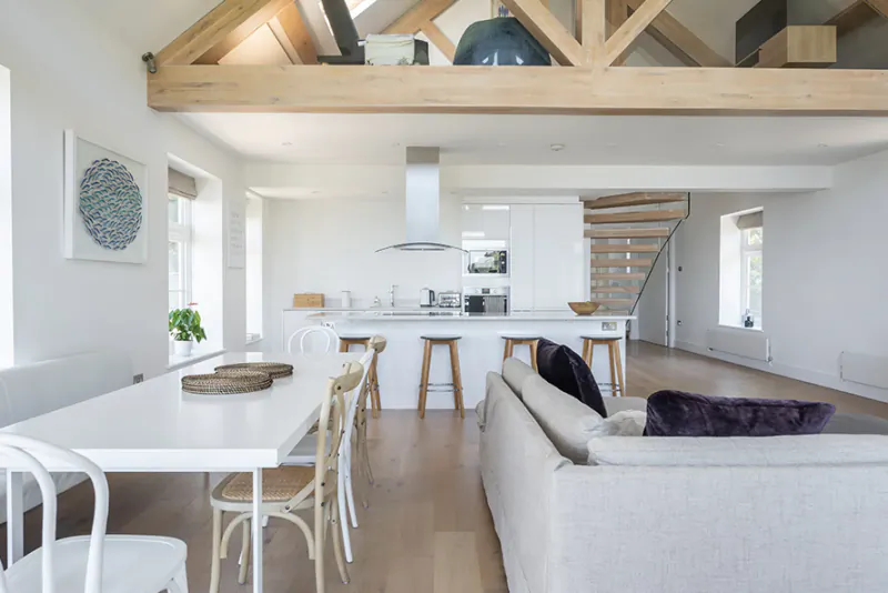 Spacious open-plan kitchen and living area with wooden beams, white island, dining table, and sofa in coastal home.