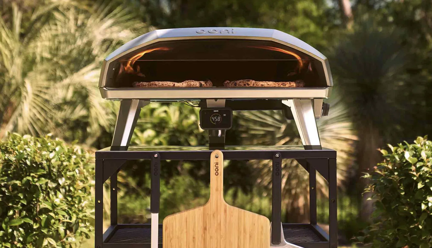Ooni pizza oven with glowing pizza inside on stand, wooden peel beside, surrounded by lush tropical plants