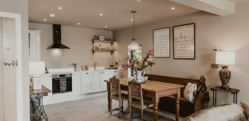 Cozy modern kitchen and dining area in The Boathouse, Wales, with white cabinets, wooden table, flowers, and lamps.