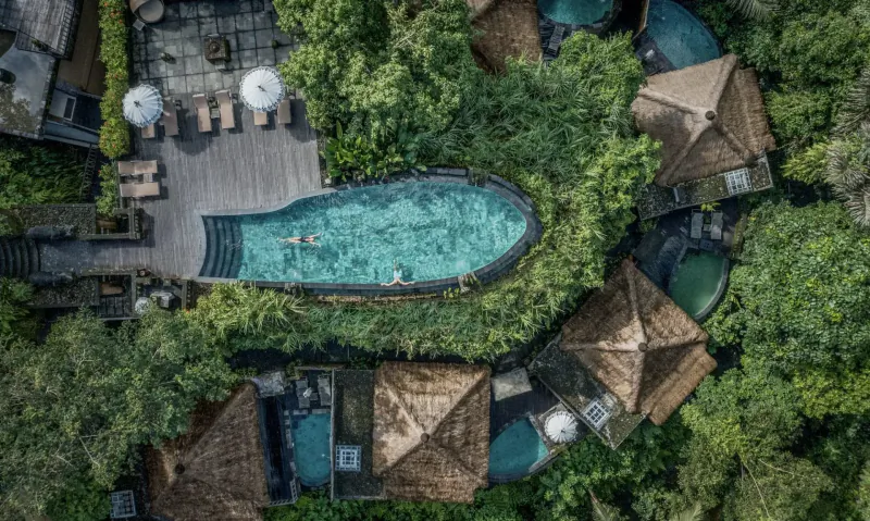 Aerial view of Kayon Resort Bali: infinity pool with swimmer, thatched villas, lush jungle surroundings.