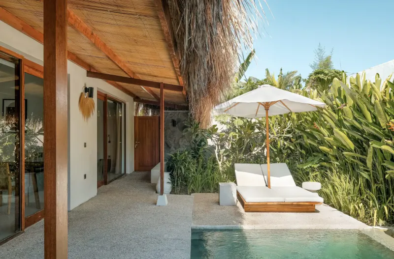 Luxury beach resort villa with thatched roof, white sun loungers by infinity pool, lush tropical garden, and blue sky.