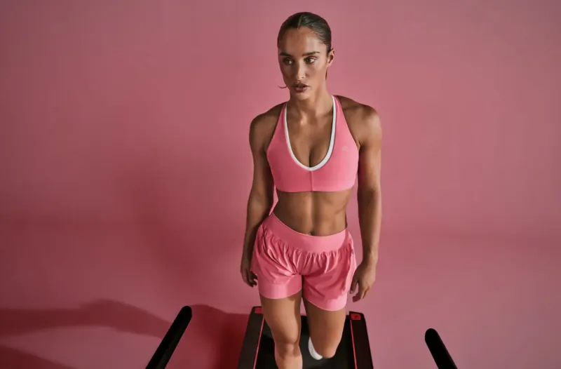 Athletic woman in pink sports bra and shorts stands on red treadmill against pink background
