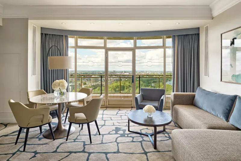 Elegant hotel living room with round dining table, blue sofa, lamps, and panoramic city park view through curved windows.