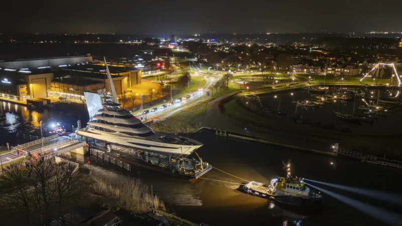 Night aerial view of nighttime construction: towering yellow stepped building beside river, tugboat maneuvering it into position amid city lights.