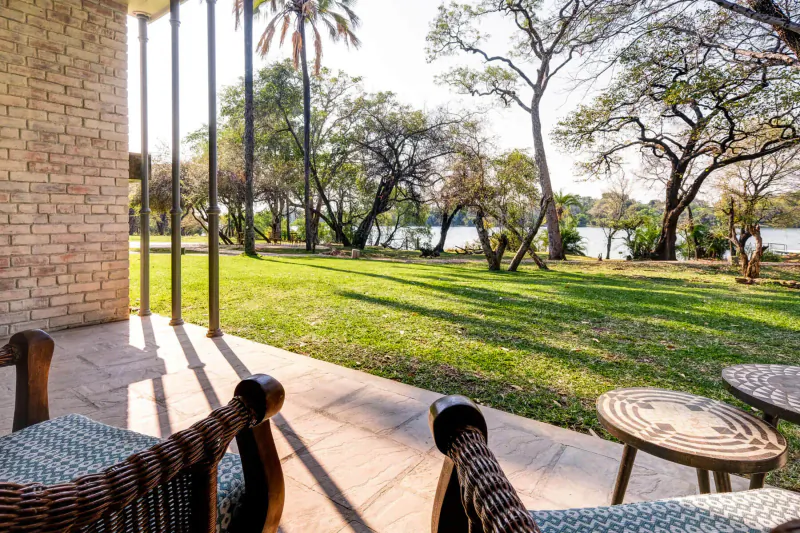 Outdoor patio with wicker chairs and table overlooking lush green lawn, palm trees, and serene river near Victoria Falls.