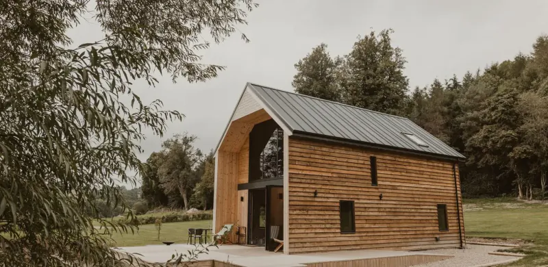 The Boathouse, a wooden cabin with arched windows beside a private lake in Wales, surrounded by trees and fields.