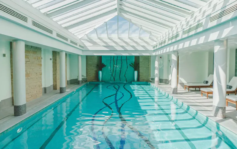Elegant indoor pool at luxury spa resort with glass ceiling, swirling wall fountain, and lounge chairs.