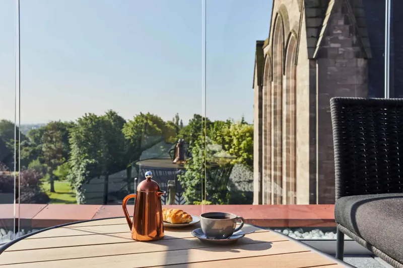 Outdoor table at Hotel Indigo Chester with copper coffee pot, croissant, and cup, overlooking historic church and green landscape.