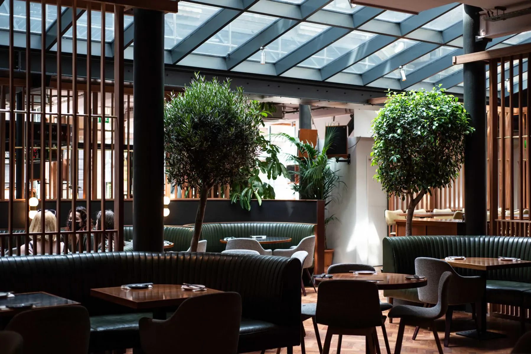 Elegant Tokyo-inspired tavern interior with green velvet seating, wooden tables, potted trees, and glass atrium ceiling.