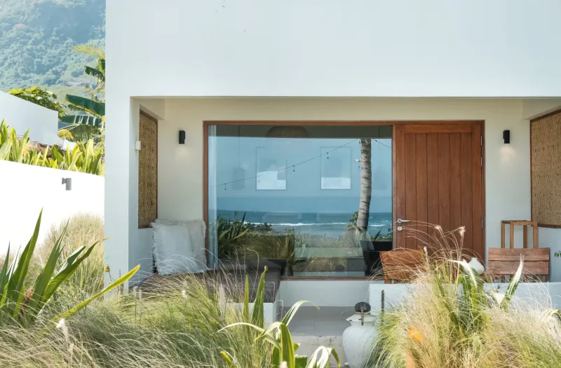 Modern white villa at Amber Lombok Beach Resort with large glass doors opening to ocean view, surrounded by tropical plants and beach grass.