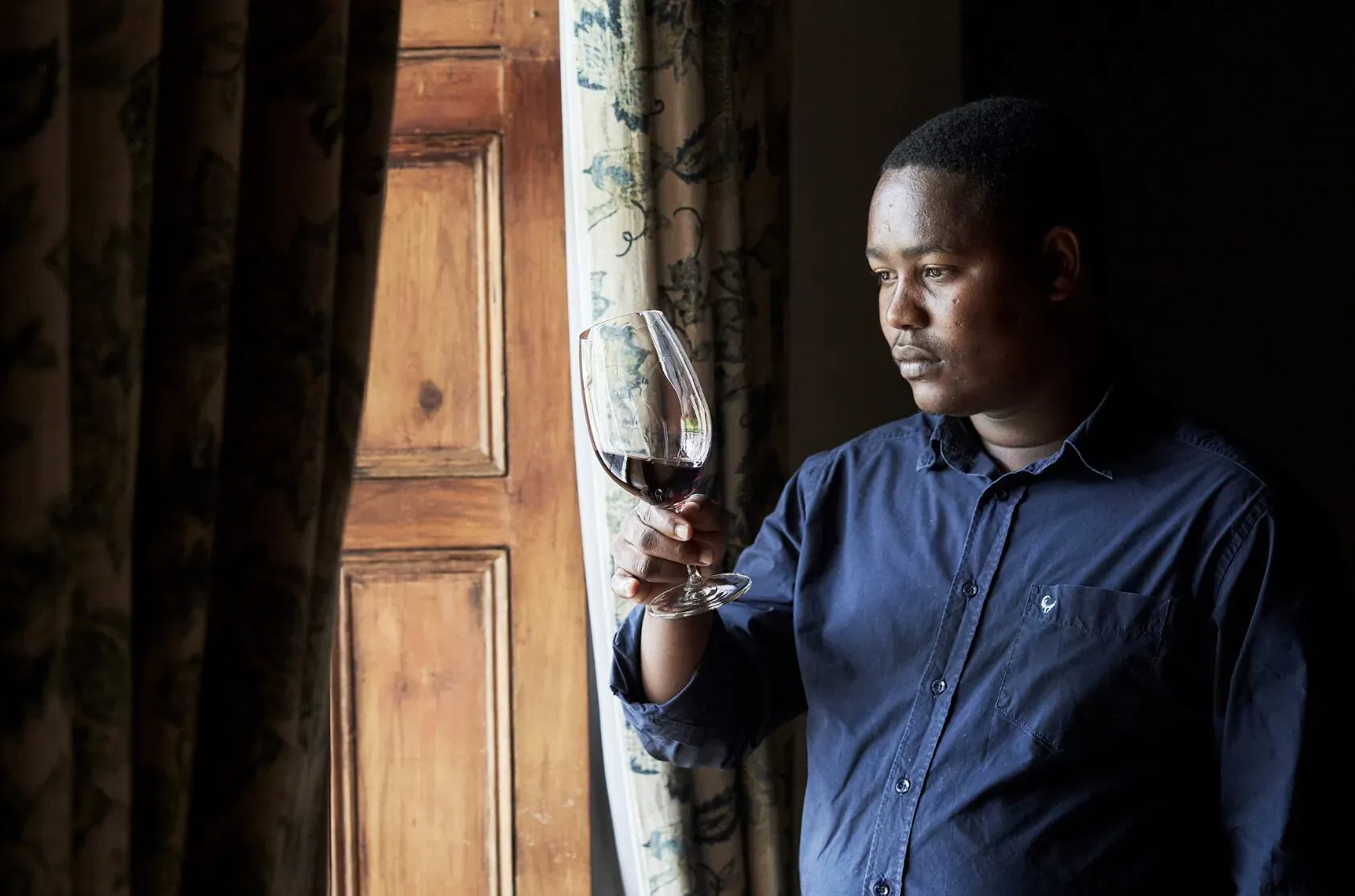 Vusi Dalicuba, Vergenoegd Löw winemaker, holds a glass of red wine, gazing out a curtained wooden door.