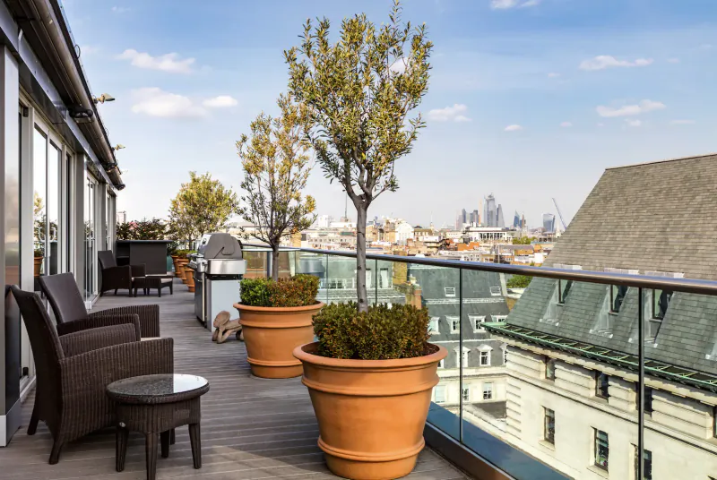 Spacious rooftop terrace at The May Fair Hotel with lounge chairs, potted trees, BBQ, and city skyline view.