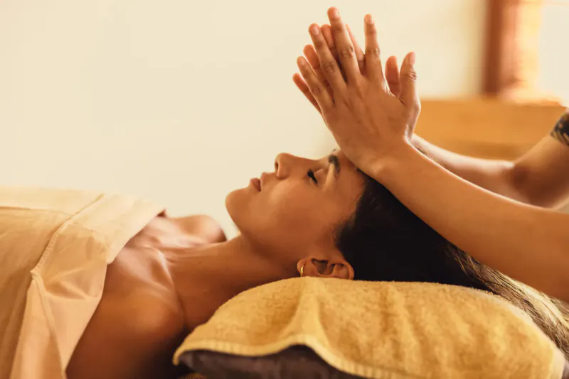 Spa therapist performing head massage on relaxed woman lying on treatment bed at resort.
