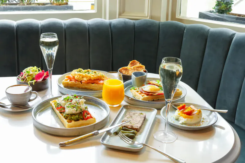 Luxury hotel table with avocado toast, eggs, sandwiches, champagne, coffee, and juice on elegant blue booth by window.