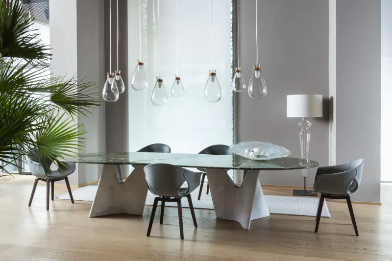 Modern glass dining table with grey curved chairs, teardrop pendant lights, palm plants in luxury grey interior.