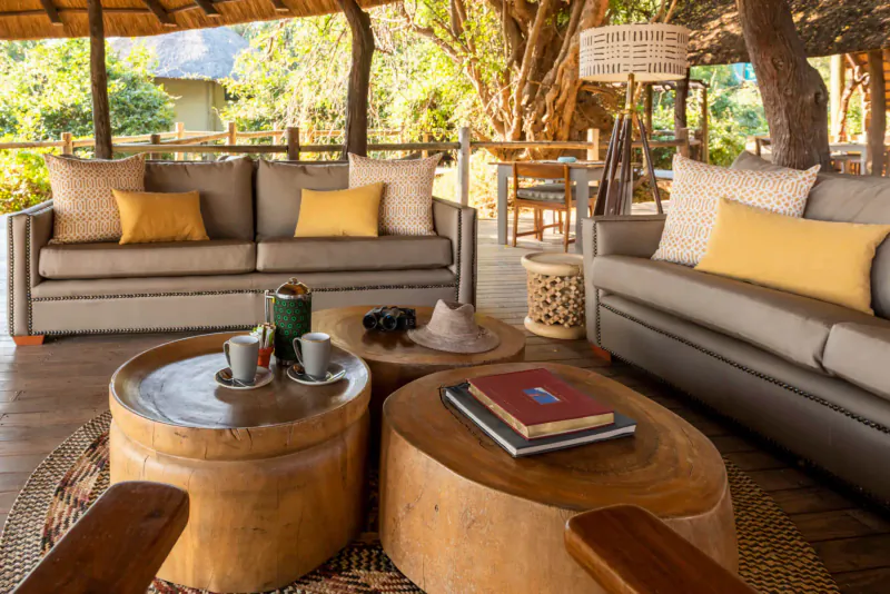 Lion Camp's outdoor lounge with beige sofas, yellow pillows, wooden tables, cups, book, and mountain backdrop under trees.