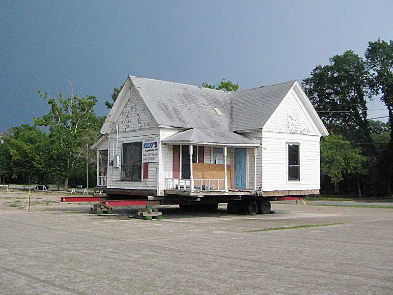 White clapboard house on a flatbed trailer truck, ready for transport on a parking lot amid trees.
