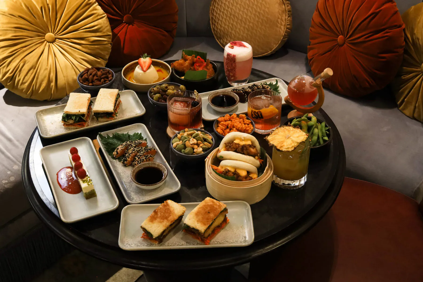 Luxurious assortment of Asian fusion dishes and cocktails on black table with plush cushions at 100 Queen's Gate Hotel