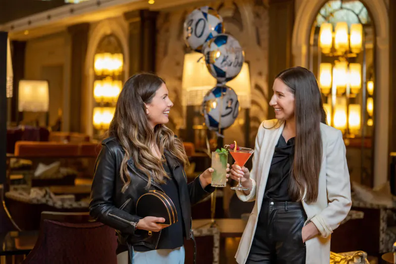 Two stylish women smiling and toasting with cocktails amid blue balloons in elegant Sheraton Grand hotel gallery