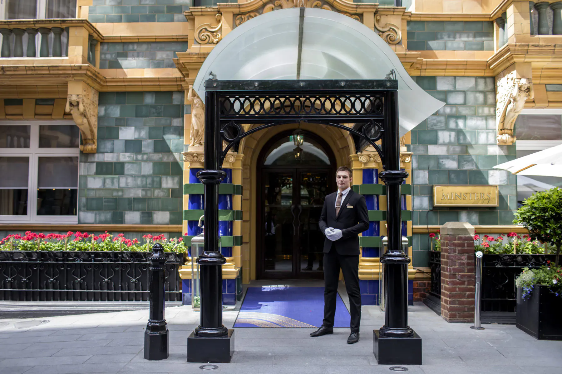 Doorman in suit stands at ornate entrance of Taj 51 Buckingham Gate Suites and Residences with flowers and blue carpet.