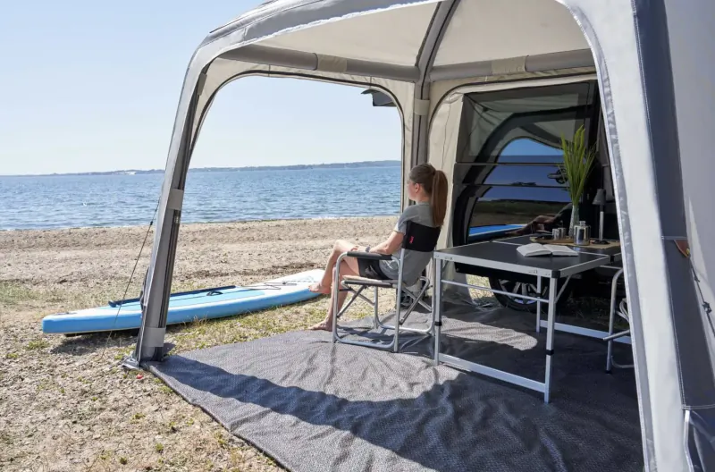 Woman sits at table in inflatable beach tent by sea, blue SUP board nearby, luxury campervan accessory.