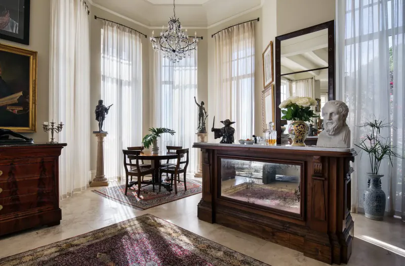 Elegant sunlit gallery room at 21 Nettleton with chandelier, marble busts, statues, round table, and large windows.
