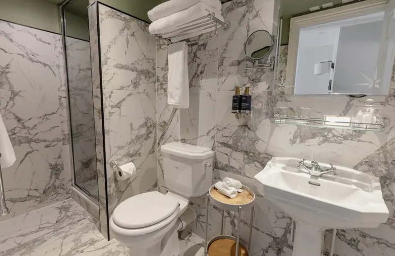 Elegant marble bathroom with glass shower, white toilet, pedestal sink, stacked towels, and toiletries.