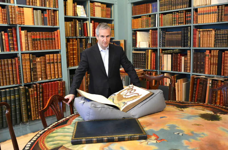 Bernard Shapero, rare book expert, in suit holding open antique book on round table in library lined with bookshelves