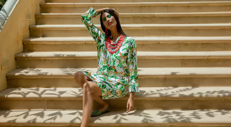 Woman in green floral long-sleeve dress and red necklace sits on beige stone stairs, one arm raised