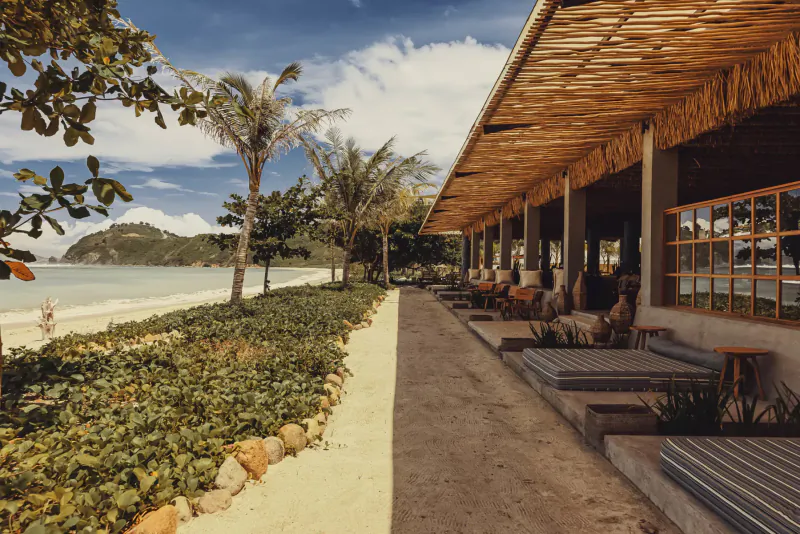 Open-air beachfront resort with thatched roof veranda, lounge seating, palm trees, and ocean view at Amber Lombok.