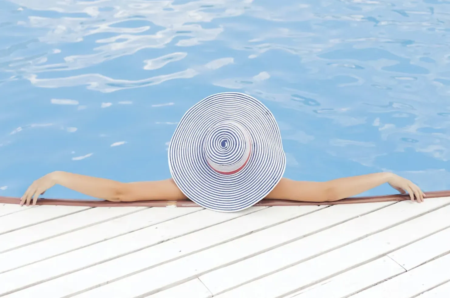 Woman in wide-brim hat relaxes with arms outstretched on pool edge, viewed from behind