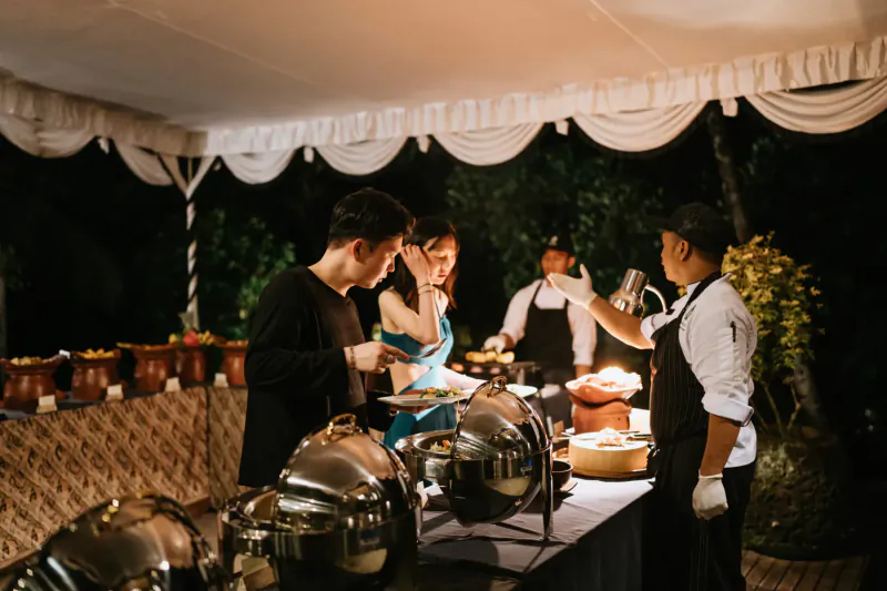 Chefs in uniforms serve food from chafing dishes to smiling guests at Kepitu Restaurant buffet under tented canopy.