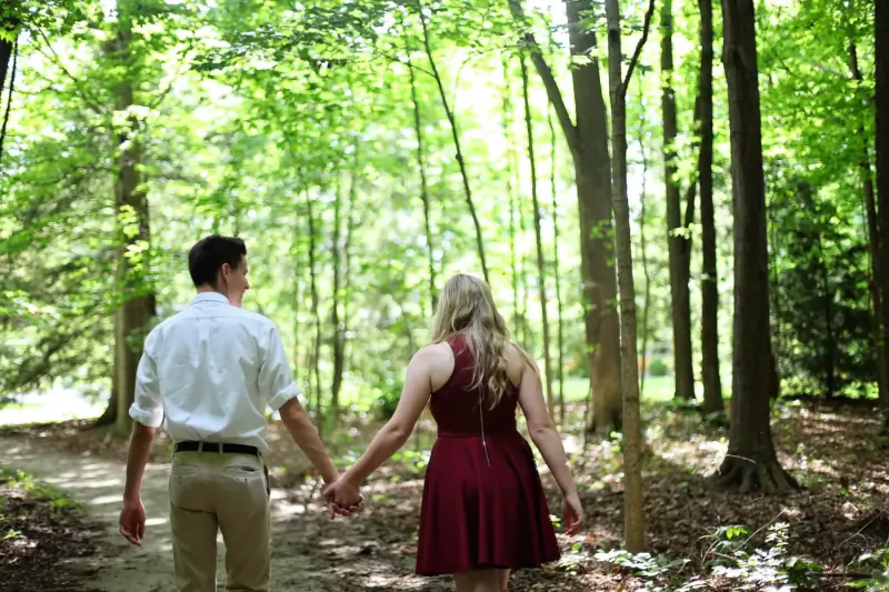 Couple holding hands walking down wooded path in lush green forest, man in white shirt and khakis, woman in red dress.
