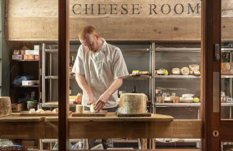 Red-haired chef in white uniform cutting wheel of cheese on wooden board in 'CHEESE ROOM', shelves of cheeses behind glass door