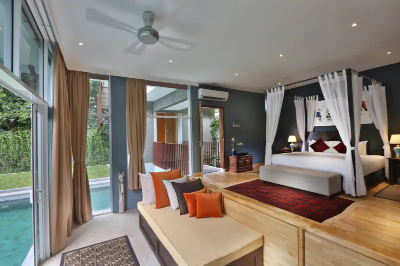 Spacious modern bedroom in wellness retreat with four-poster bed, orange pillows, pool view, and tropical garden.