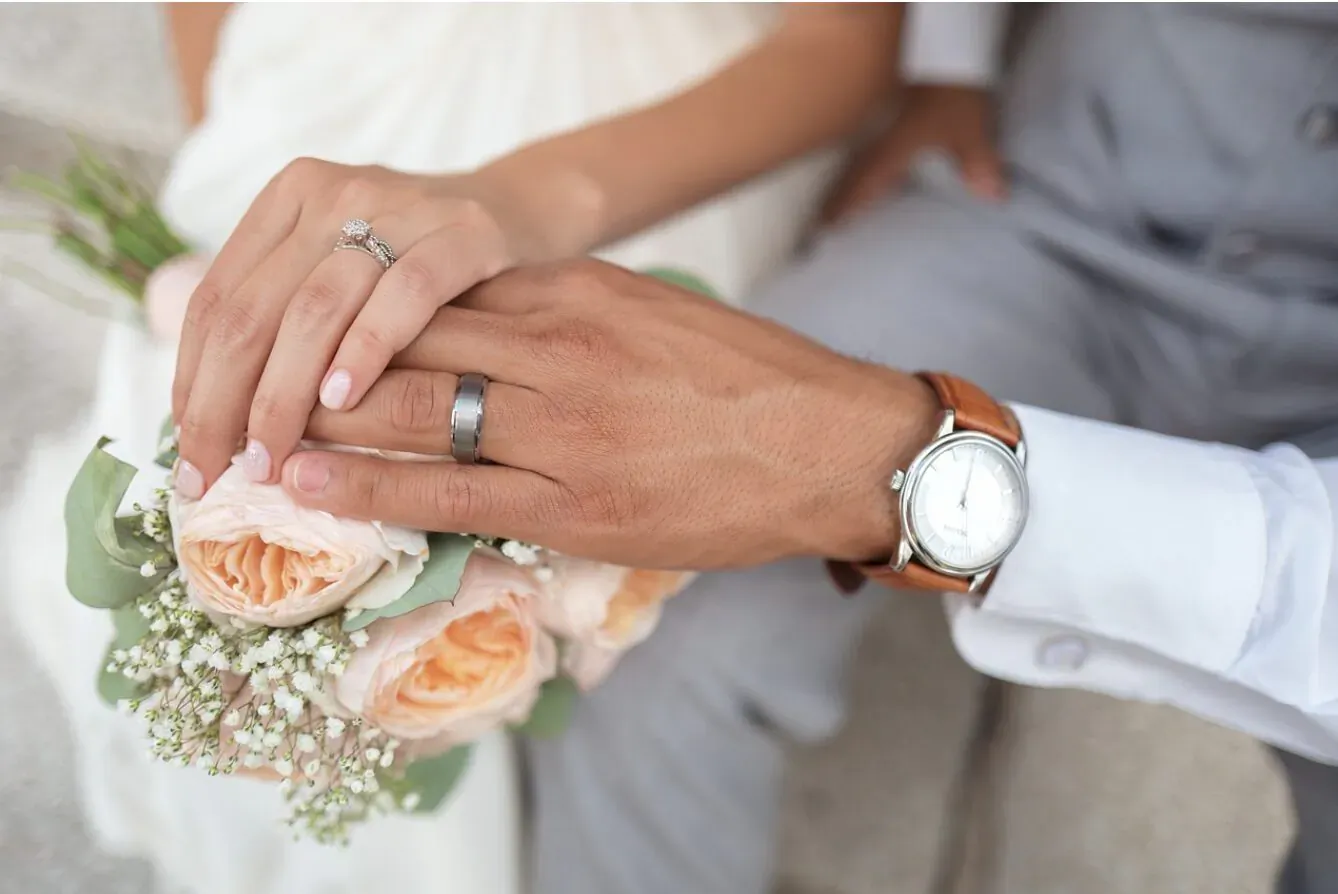 Bride and groom's hands intertwined holding peach roses bouquet, showing diamond and black tungsten wedding bands and watch