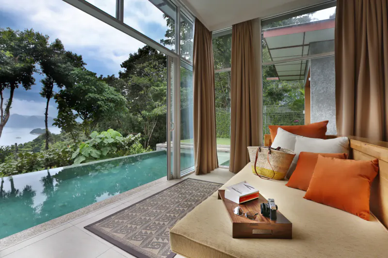 Luxurious bedroom with orange cushions and tray on low bed, floor-to-ceiling windows overlooking lush jungle, infinity pool, and sea.