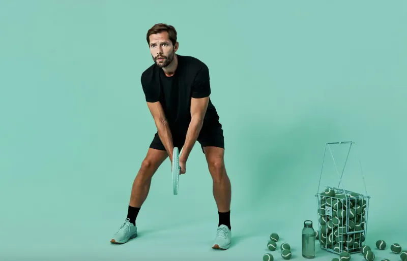 Athletic man in black athletic wear poised with racket on mint green background with basket of tennis balls