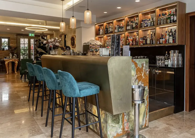 Elegant hotel bar with turquoise velvet barstools, brass counter, marble accents, and extensive liquor shelves.