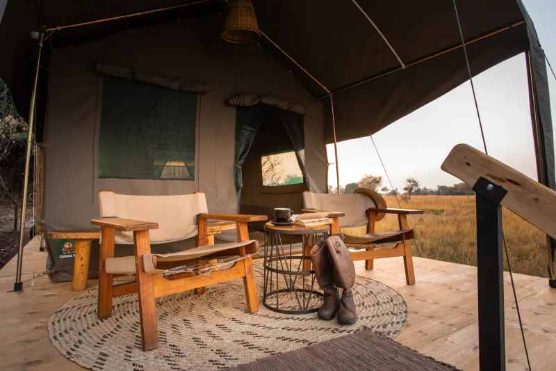 Spacious safari tent deck with wooden chairs, round rug, boots propped on table, overlooking grassy plains.