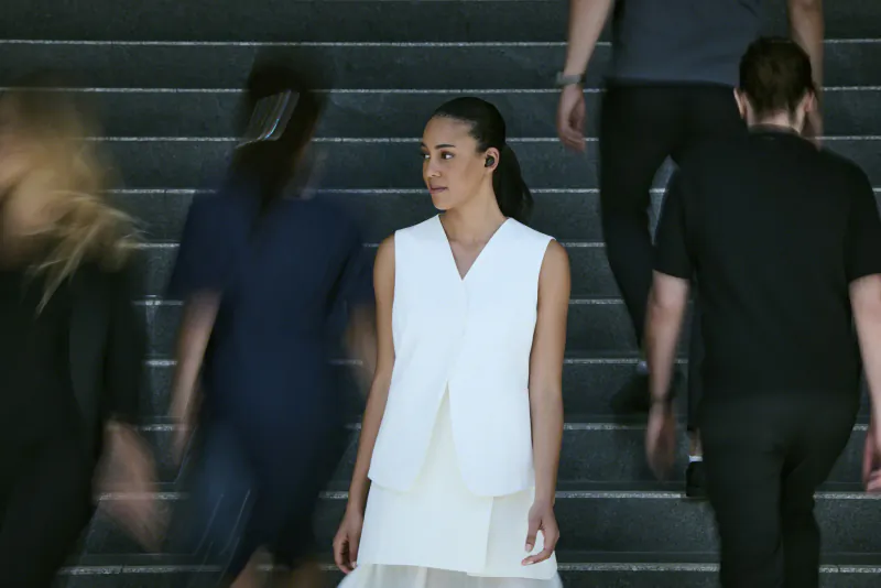 Woman in white layered dress and earbuds stands on stairs amid blurred crowd, Sony WF-1000XM6 earbuds promo