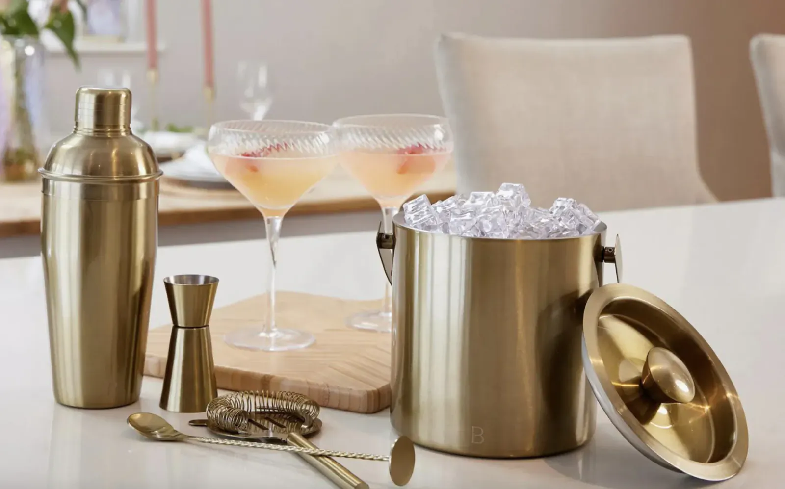 Gold cocktail shaker set with ice bucket, two pink martini glasses with fruit, bar tools on wooden board and table.
