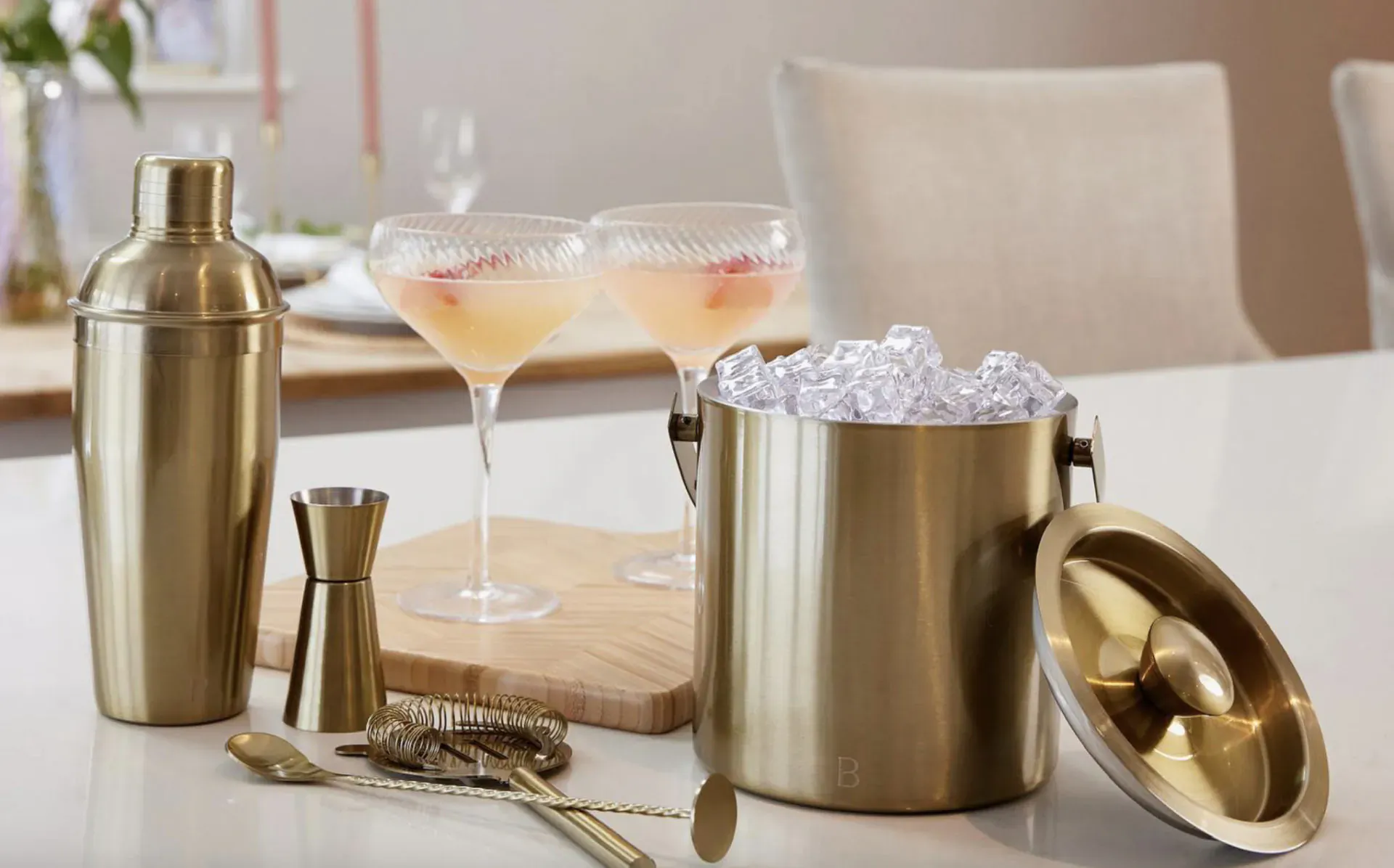 Gold cocktail shaker set with ice bucket, two pink martini glasses with fruit, bar tools on wooden board and table.