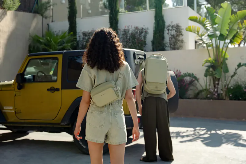 Two people in stylish green outfits and backpacks walk toward a yellow Jeep Wrangler in sunny tropical courtyard.