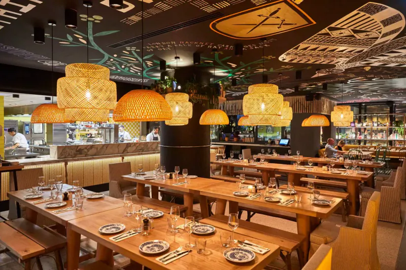 Modern restaurant interior with woven lamps, artistic ceiling murals, wooden tables set with plates and glasses, open kitchen.
