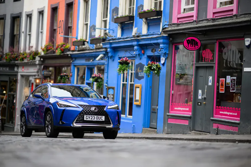 Blue Lexus UX crossover parked on wet street before colorful Georgian buildings at Virgin Hotel, Edinburgh, with pink 'PINK' shop sign.